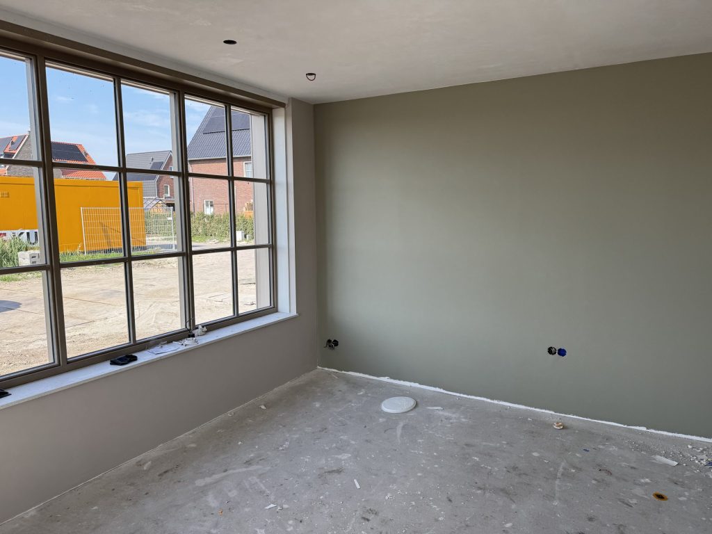 Nieuwbouwkamer met een groengrijs gesausde wand, een grote raampartij en nog een betonvloer, met buiten een bouwterrein