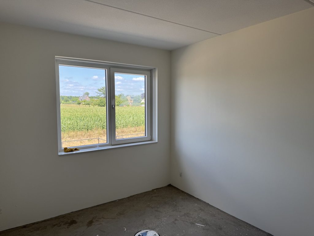 Lege slaapkamer in een nieuwbouwwoning met gladde witte wanden en een raam met uitzicht op een maisveld