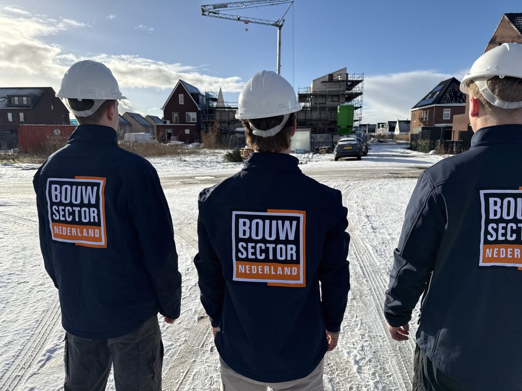 Drie vakmensen van Bouwsector Nederland in werkjassen en met witte helmen op een besneeuwde bouwweg tussen nieuwe woningen en een bouwkraan